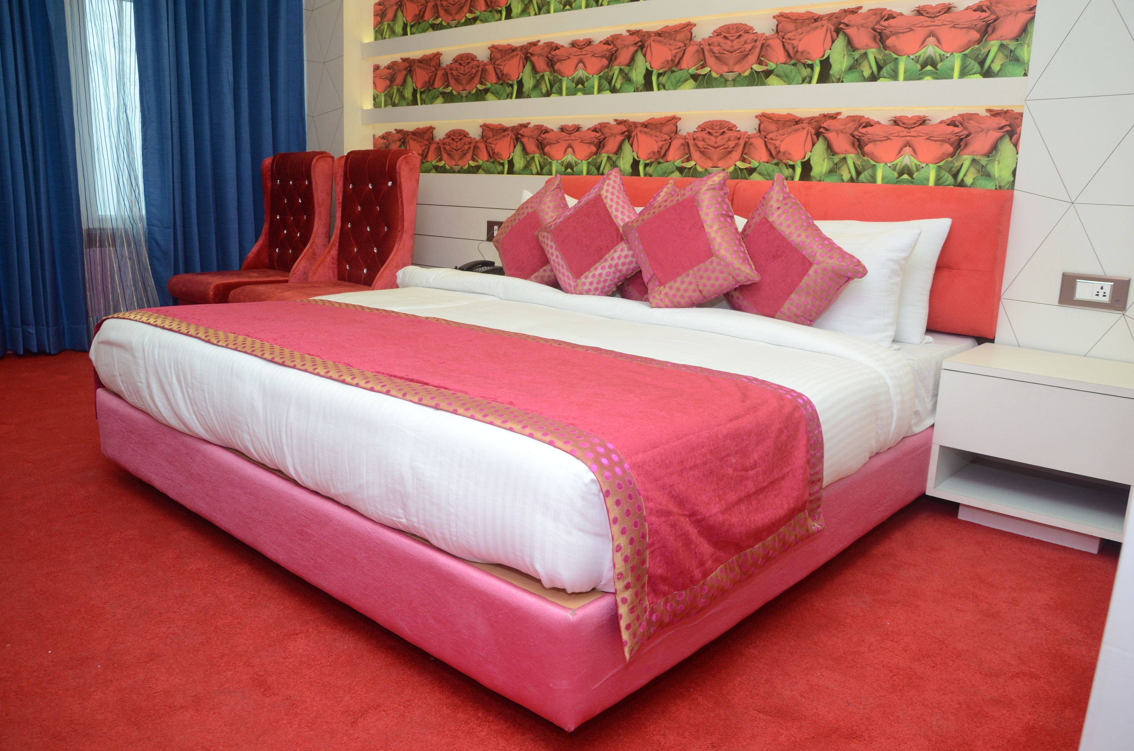Habitación de hotel con una cama grande, ropa de cama roja y blanca, una alfombra roja y una pared con estampado de rosas, además de dos sillas rojas.