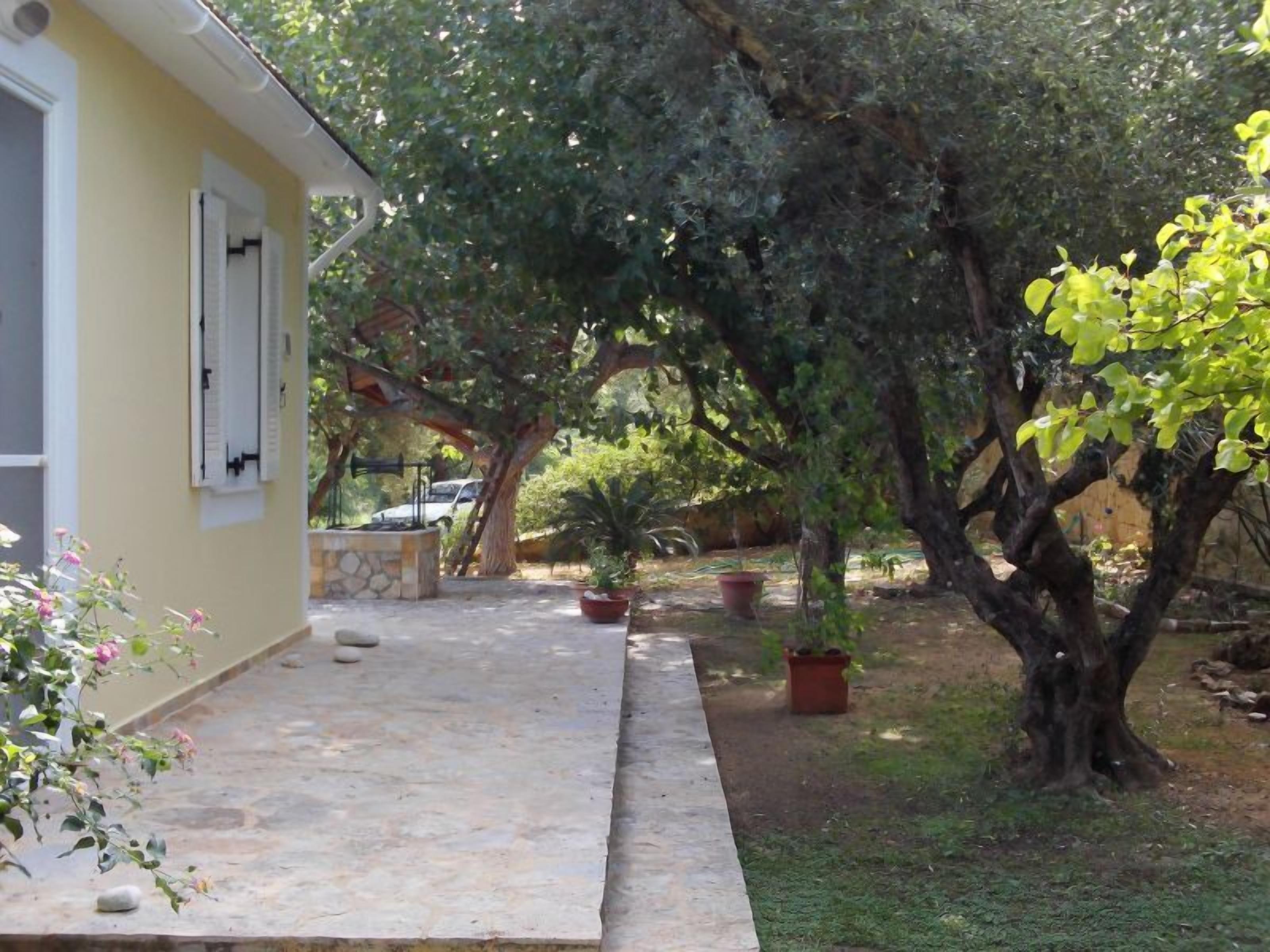 A yellow building with a stone pathway leading to a garden area with potted plants and a large tree.