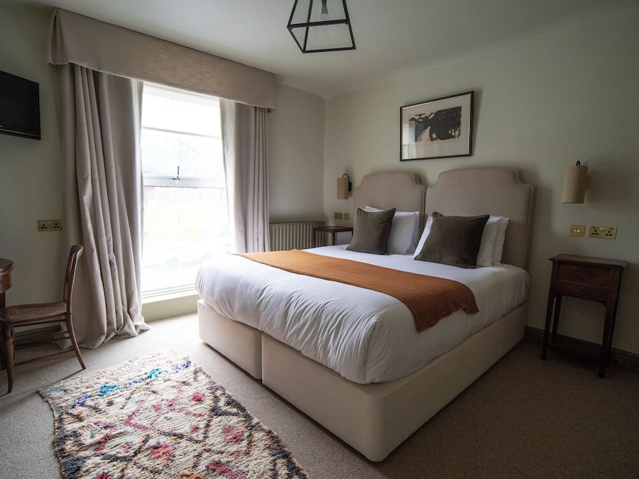 Une chambre à coucher bien rangée, avec un grand lit, une table de chevet, une chaise, un tapis et une fenêtre avec des rideaux.