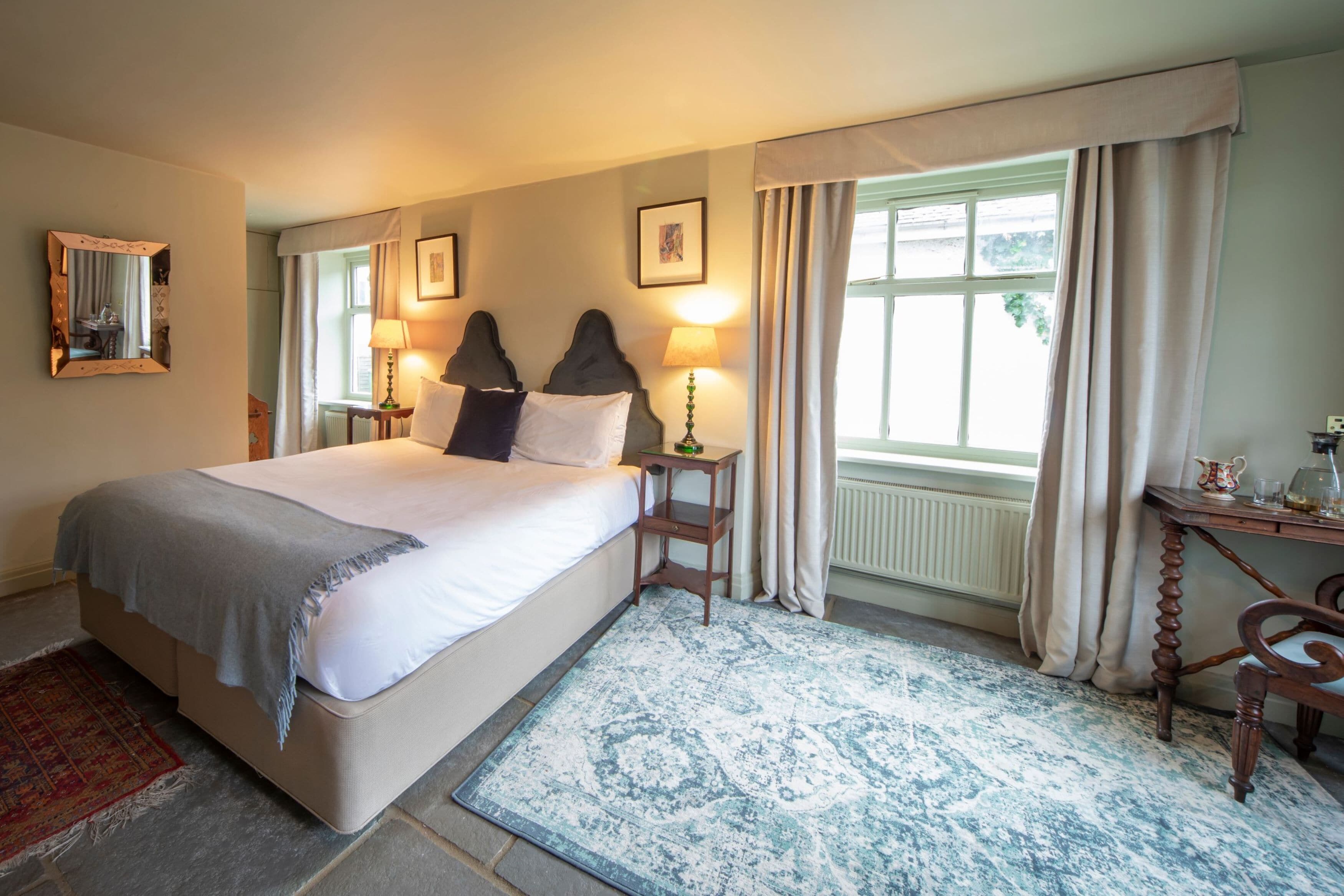 A bedroom with a large bed, two bedside lamps, a wooden nightstand, a desk with a chair, and a window with curtains.