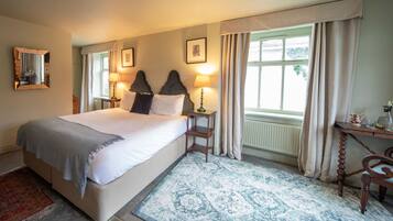 A bedroom with a large bed, two bedside lamps, a wooden nightstand, a desk with a chair, and a window with curtains.
