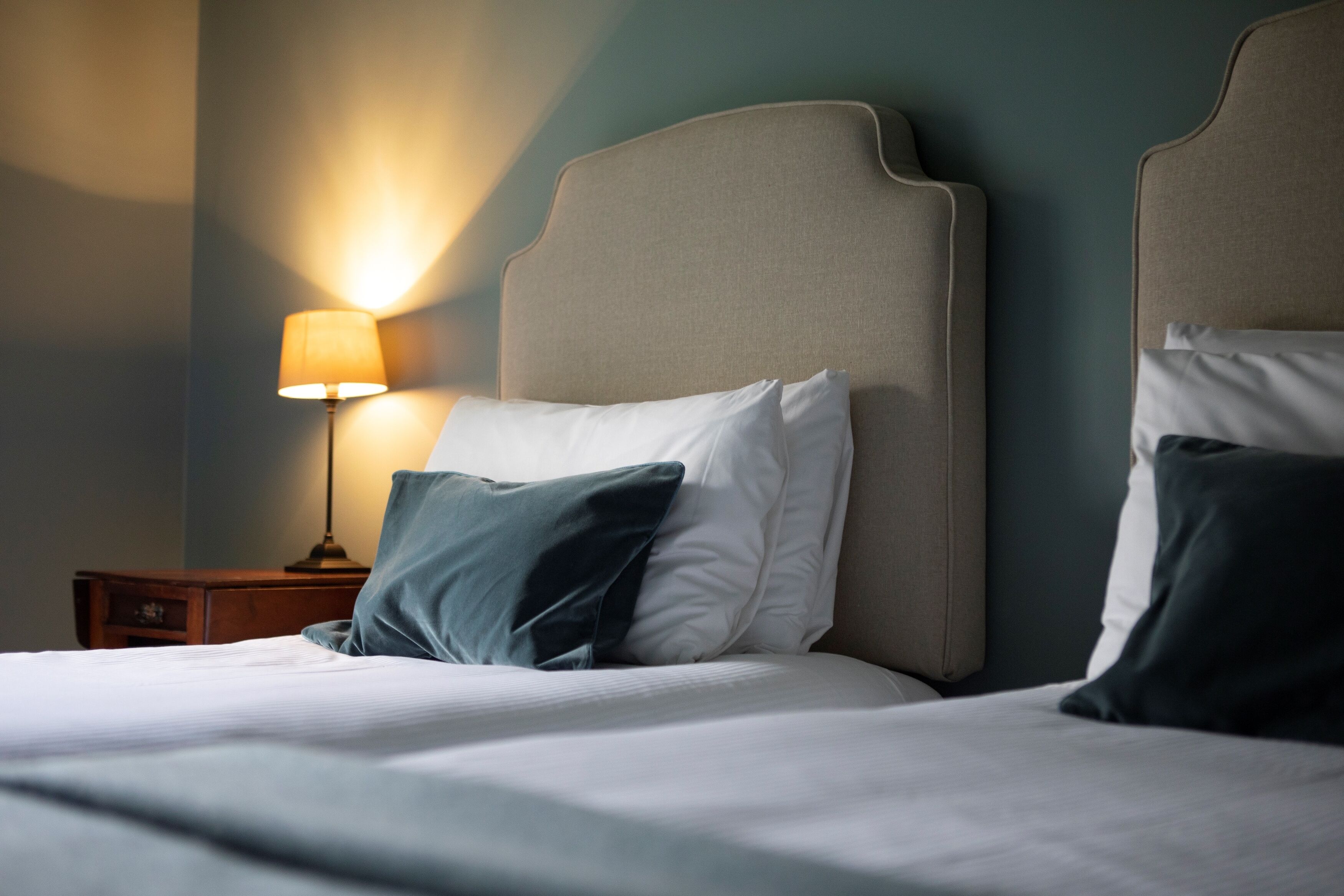 A neatly made bed with white and dark blue pillows, a wooden nightstand with a lamp, and a wall with a textured finish.