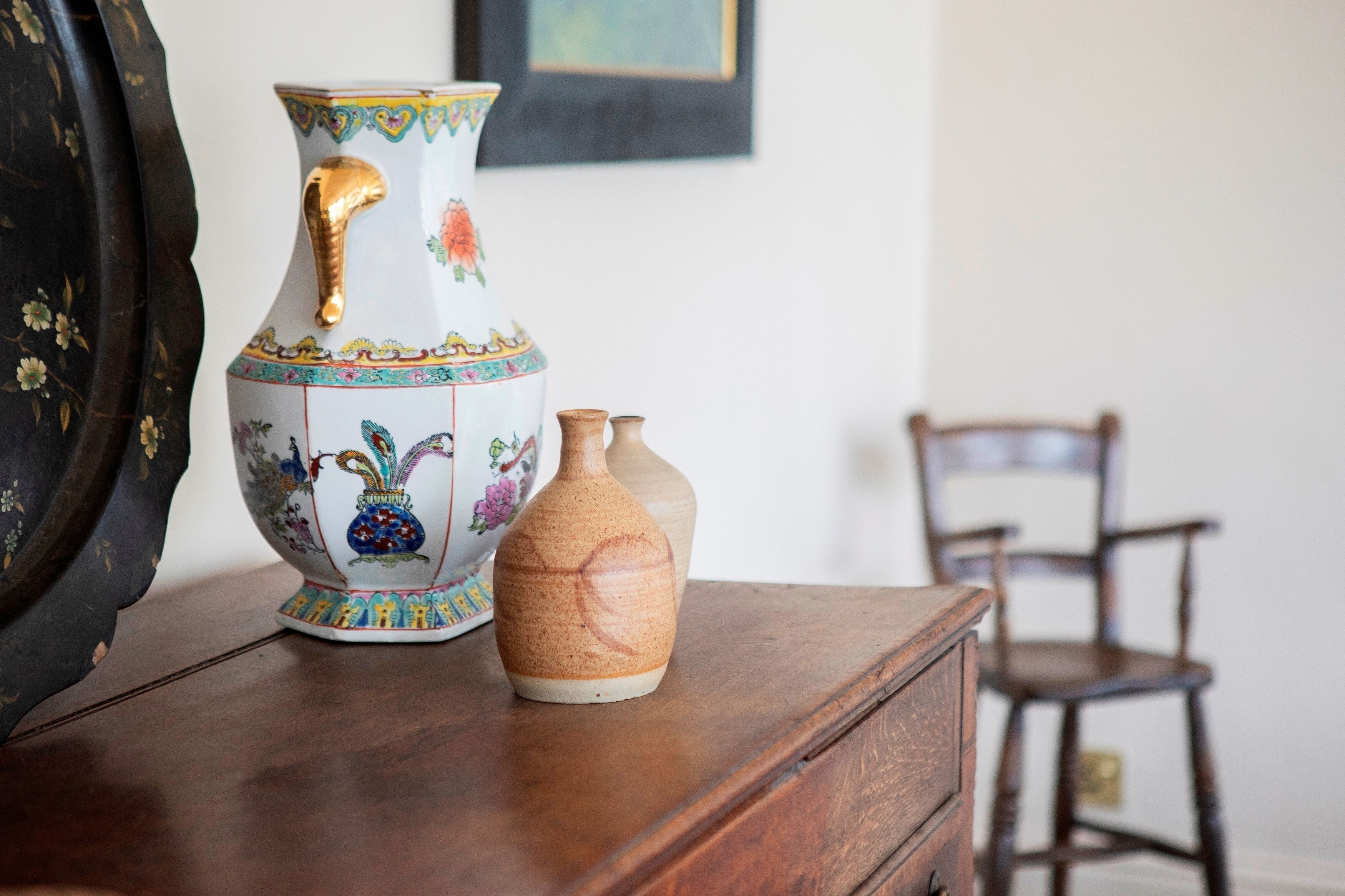 A decorative vase with floral patterns on a wooden surface, a smaller plain vase beside it, and a wooden chair in the background.