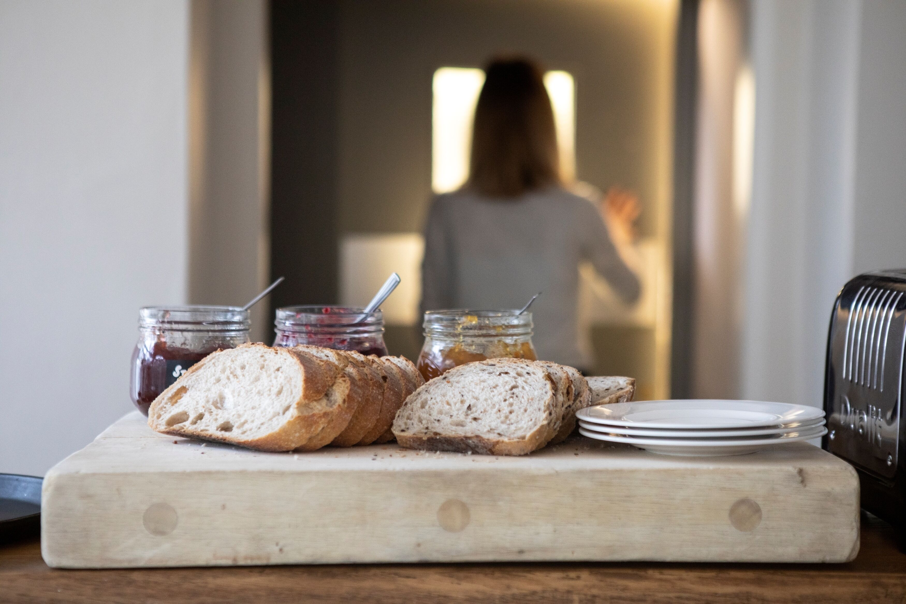 A wooden cutting board with sliced bread and jars of jam, a toaster, and plates on a kitchen counter.
