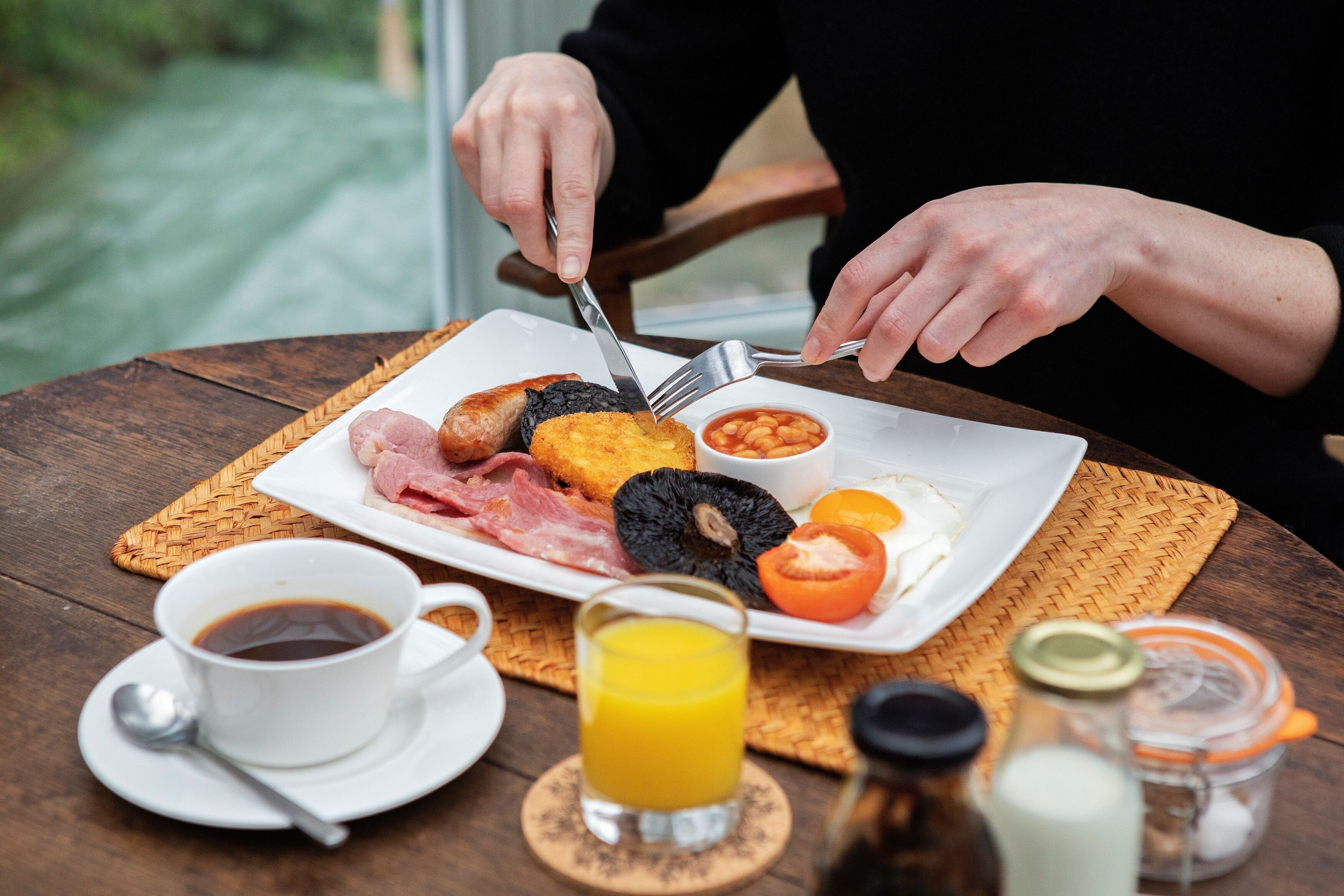 Ein Frühstücksteller mit Eiern, Speck, Blutwurst und Bohnen, eine Tasse Kaffee und ein Glas Orangensaft auf einem Holztisch.