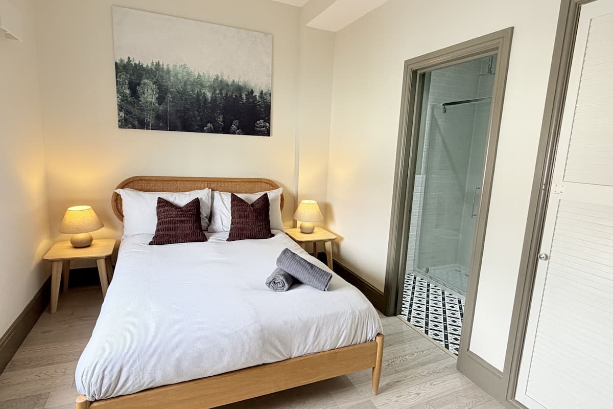 A neatly made bed with a headboard, two bedside lamps, a painting above the bed, and a door with a patterned floor mat.
