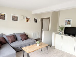 A living room with a grey sofa, a wooden coffee table, a TV on a cabinet, and framed pictures on the wall.