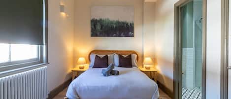 A bedroom with a bed, two bedside lamps, a painting on the wall, and a view of a bathroom through an open door.