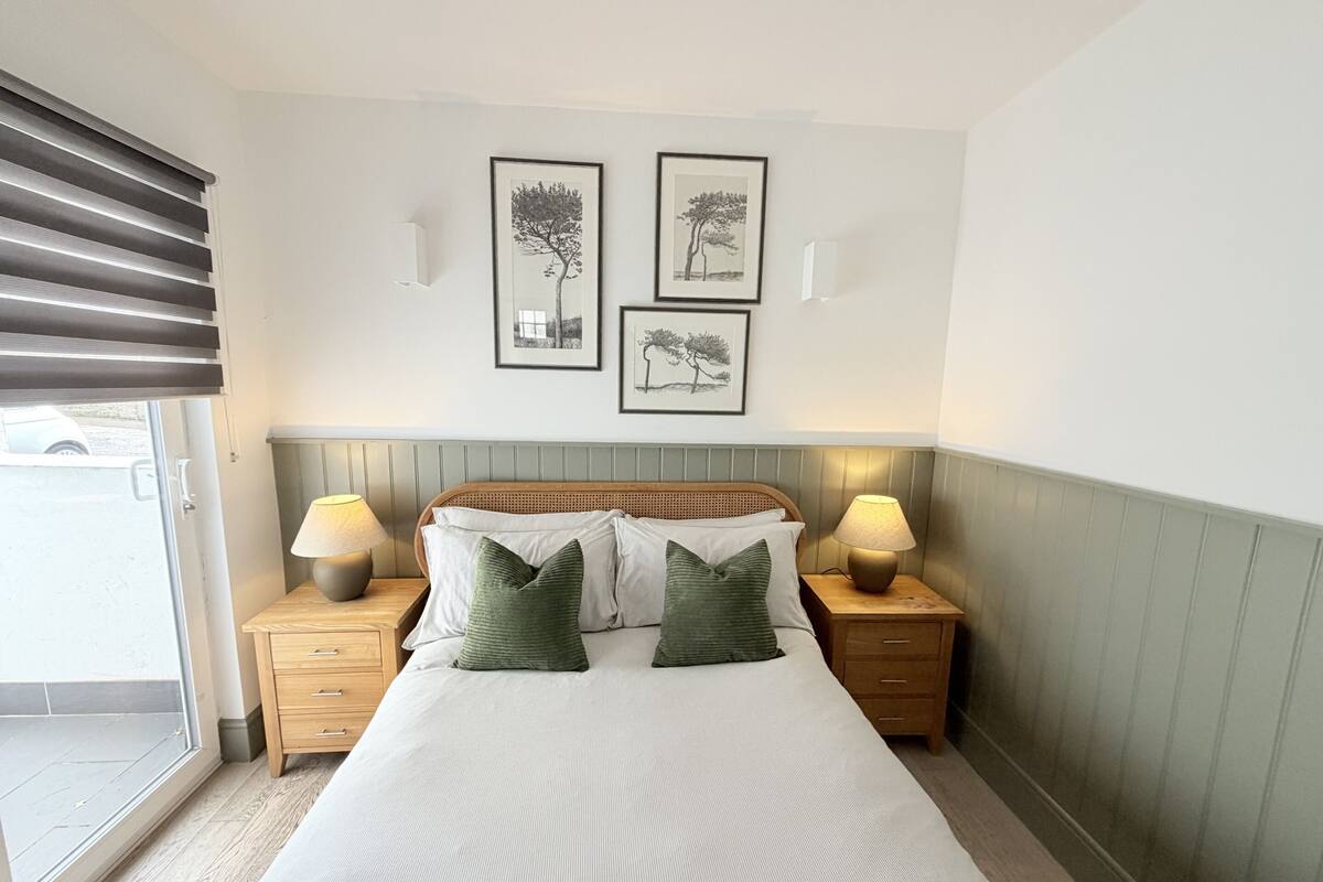 A bedroom with a bed, two bedside tables, a window with blinds, and three framed pictures on the wall.