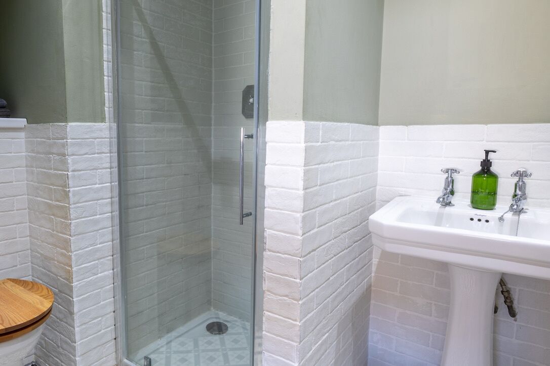 A bathroom with a glass-enclosed shower, a white sink with a green soap dispenser, and a wooden toilet.