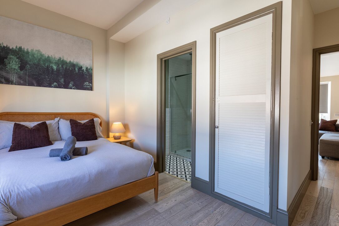 A bedroom with a bed, a nightstand, a lamp, a glass door leading to a shower, and a painting on the wall.