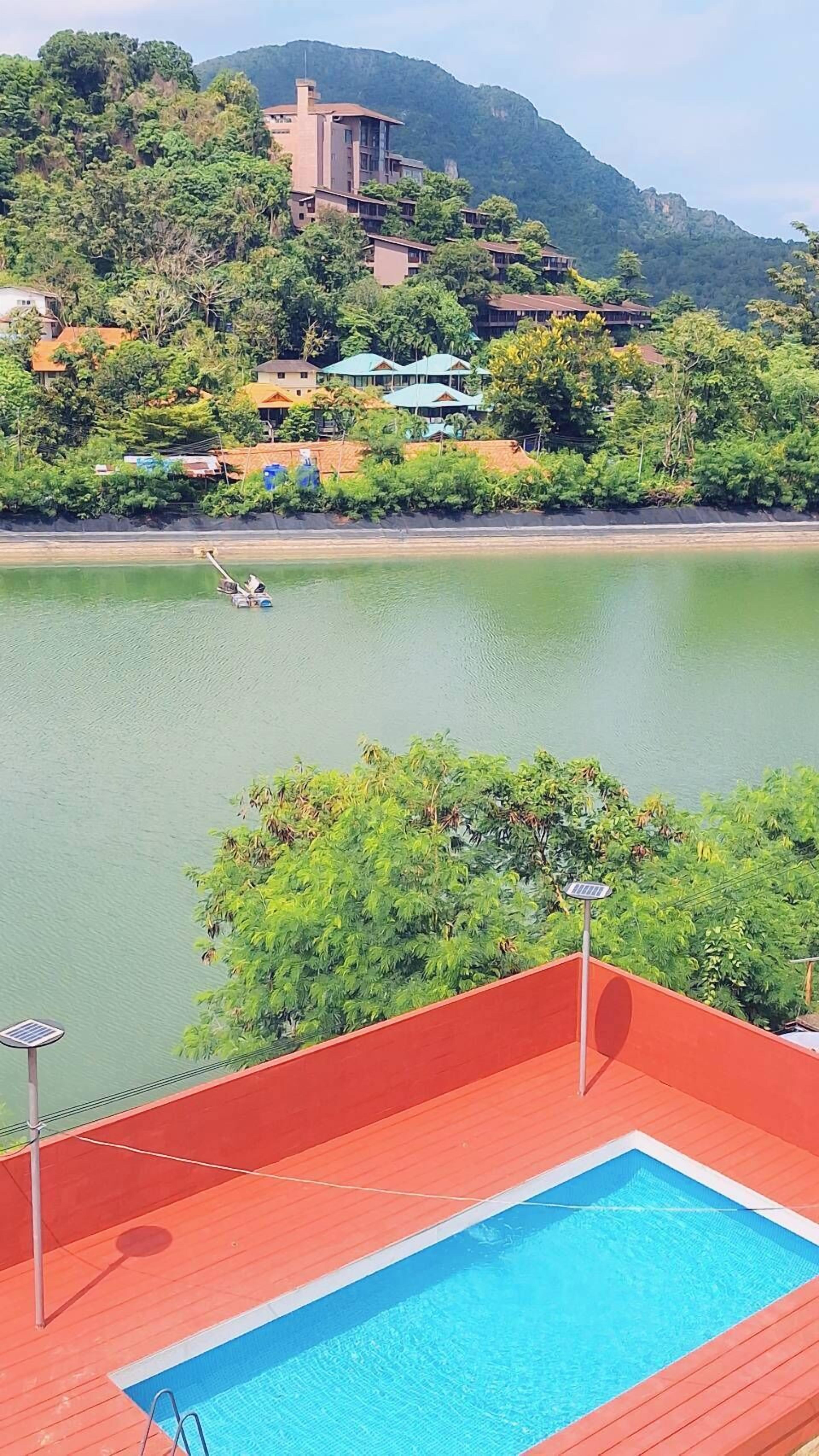 A swimming pool with a wooden deck, surrounded by lush greenery and a view of a mountain and buildings in the background.
