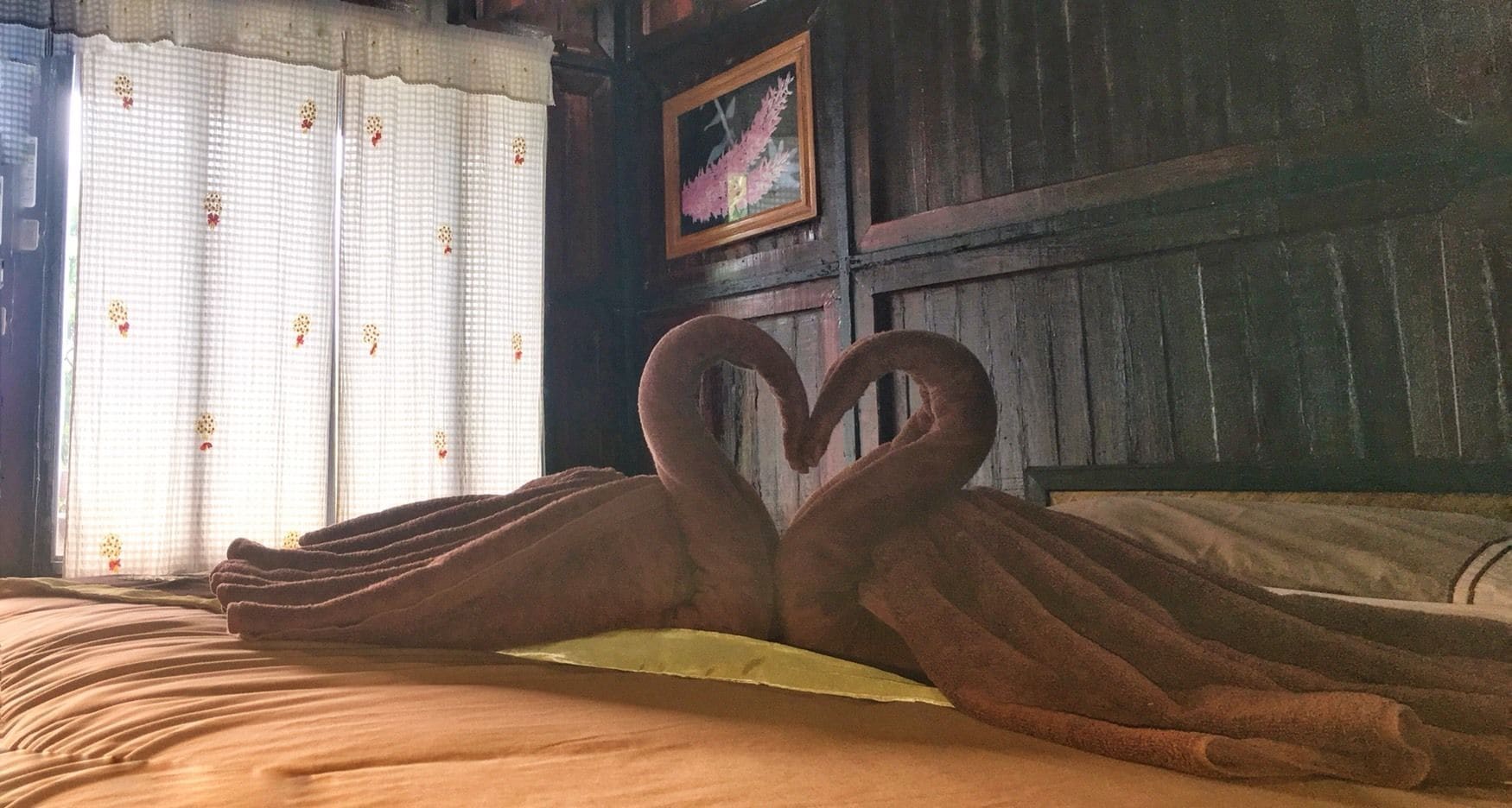 Una cama con una toalla dispuesta en forma de corazón, una ventana con cortinas y un cuadro en la pared.