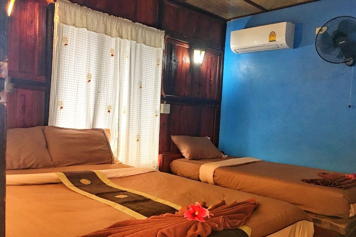 Ein Zimmer mit zwei Betten, einem fensterbelaubten Fenster, einer Klimaanlage und einem Wandventilator.