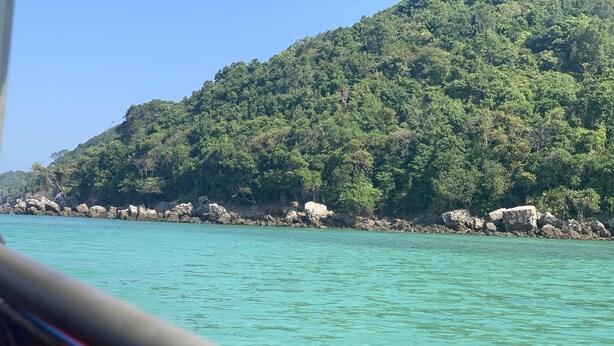 Blick auf eine Küstenlandschaft mit klarem türkisblauem Wasser und einem bewaldeten Hügel im Hintergrund.