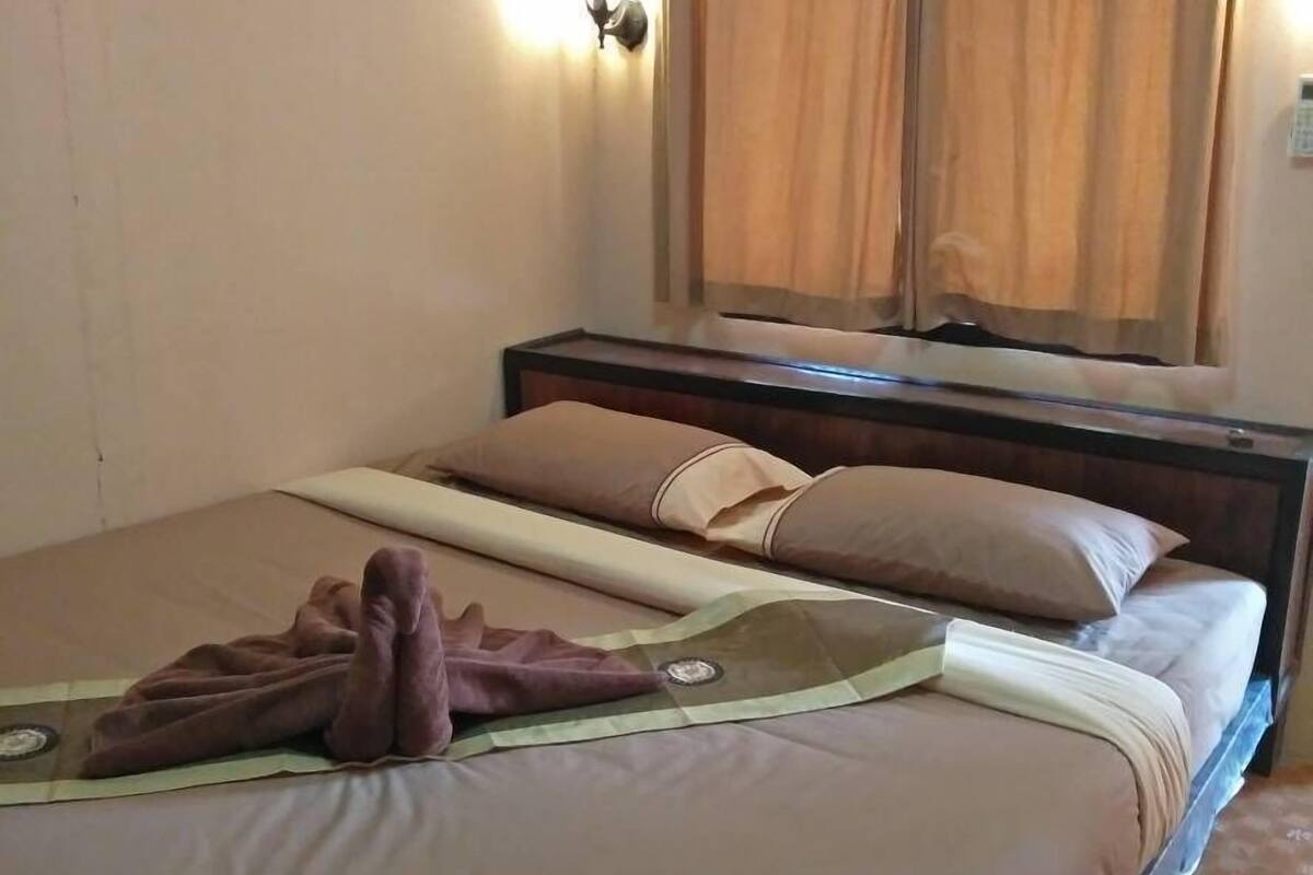 Ein ordentlich bezogenes Bett mit einer braunen Decke und einem gefalteten Handtuch, ein Holz-Kopfteil und ein Fenster mit Vorhängen.