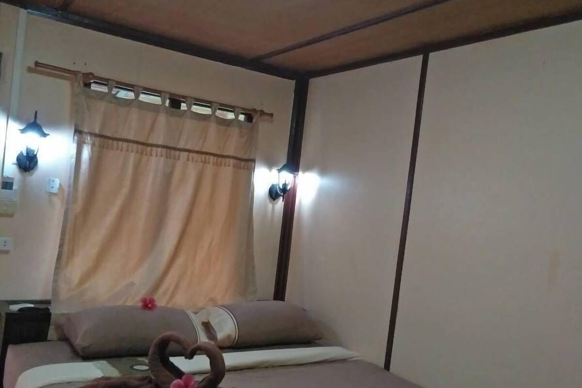 Ein Einzelbett mit brauner Decke, einem dekorativen Schwanenkissen und einem kissenförmigen Schwan. Ein Fenster mit Vorhang und Wandleuchten sind vorhanden.