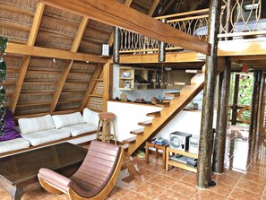 Ein großzügiger Wohnbereich mit hohen Decken, Holzbalken, einer Couch, einem gepolsterten Sessel, einem kleinen Tisch mit elektronischen Geräten und einer Küchenzeile im Hintergrund.