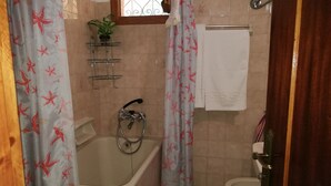 Un baño con bañera, ducha, inodoro y lavamanos. Hay cortinas con estampado de estrella de mar, un perchero y un estante pequeño con una planta.