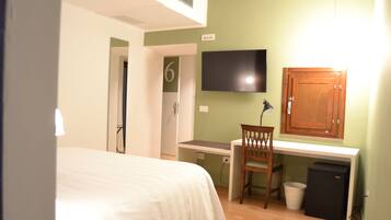 Una habitación de hotel con cama, escritorio, silla, televisor y un pequeño cubo de basura.