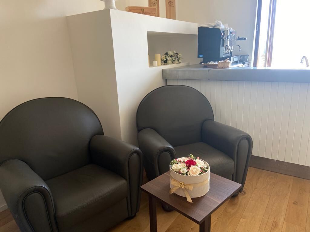 Ett rum med två mörka läderfåtöljer, ett litet träbord med en dekorativ blomsteruppsättning och ett litet kök i bakgrunden.