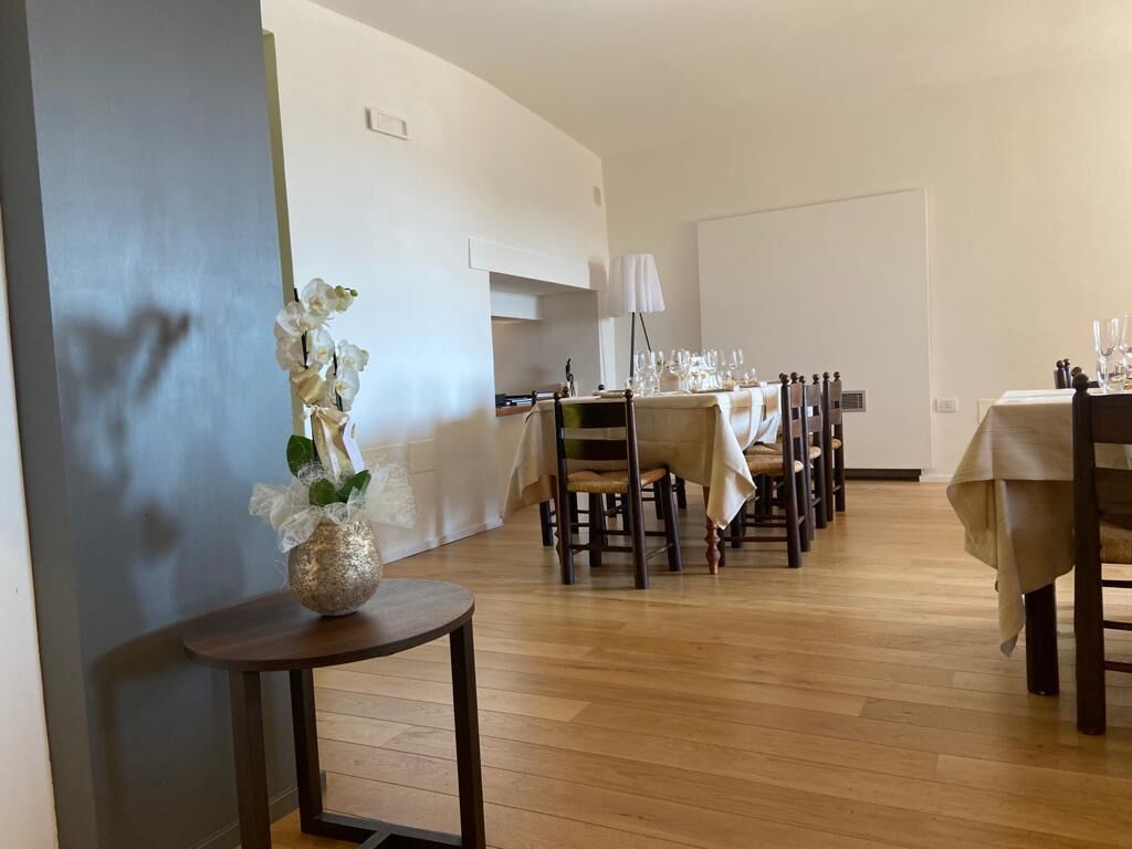 Un comedor con suelo de madera, varias mesas dispuestas para comer y un jarrón con flores blancas sobre una mesita auxiliar.
