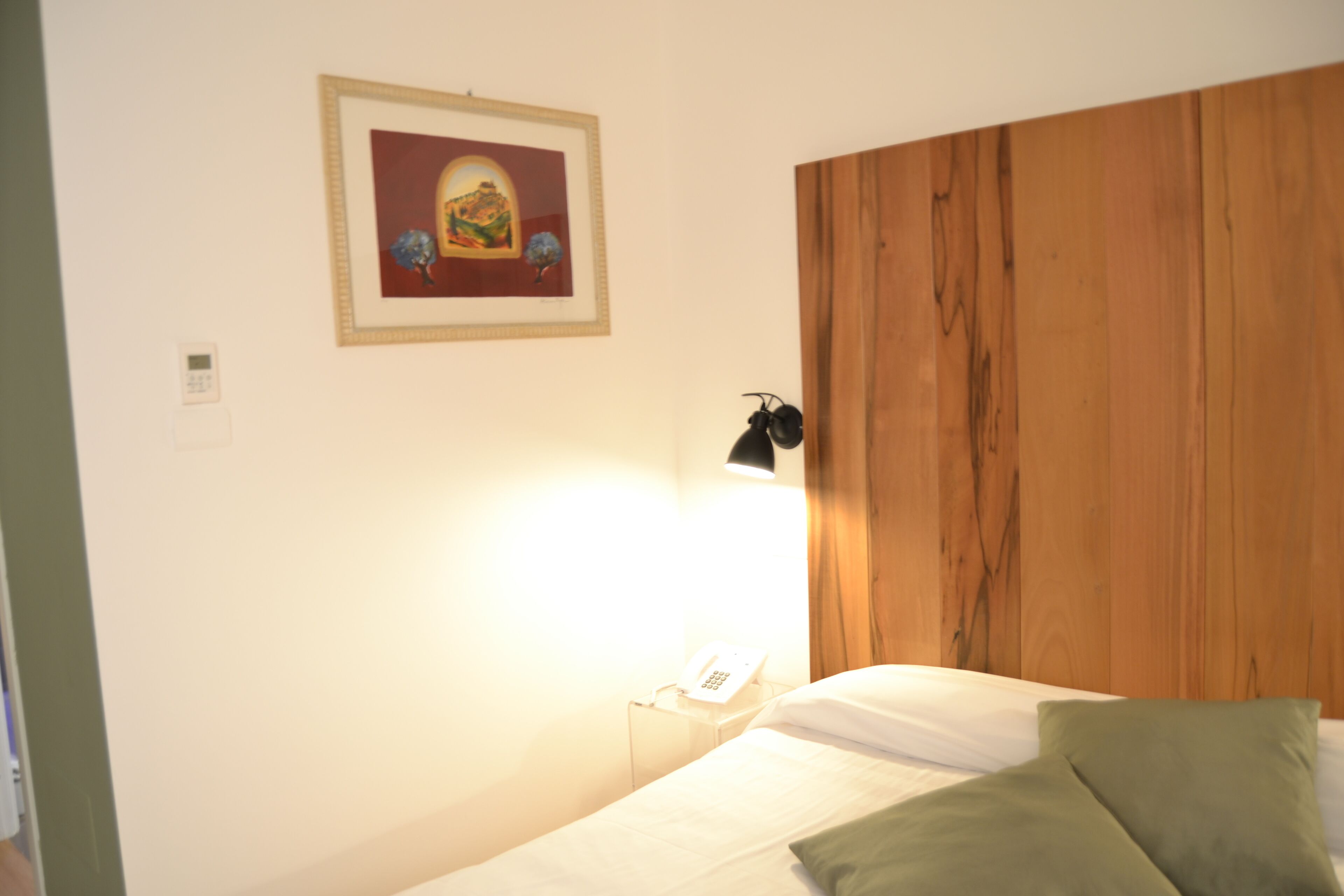 A hotel room with a wooden headboard, a bed with white linens, a green pillow, a framed picture on the wall, and a bedside lamp.