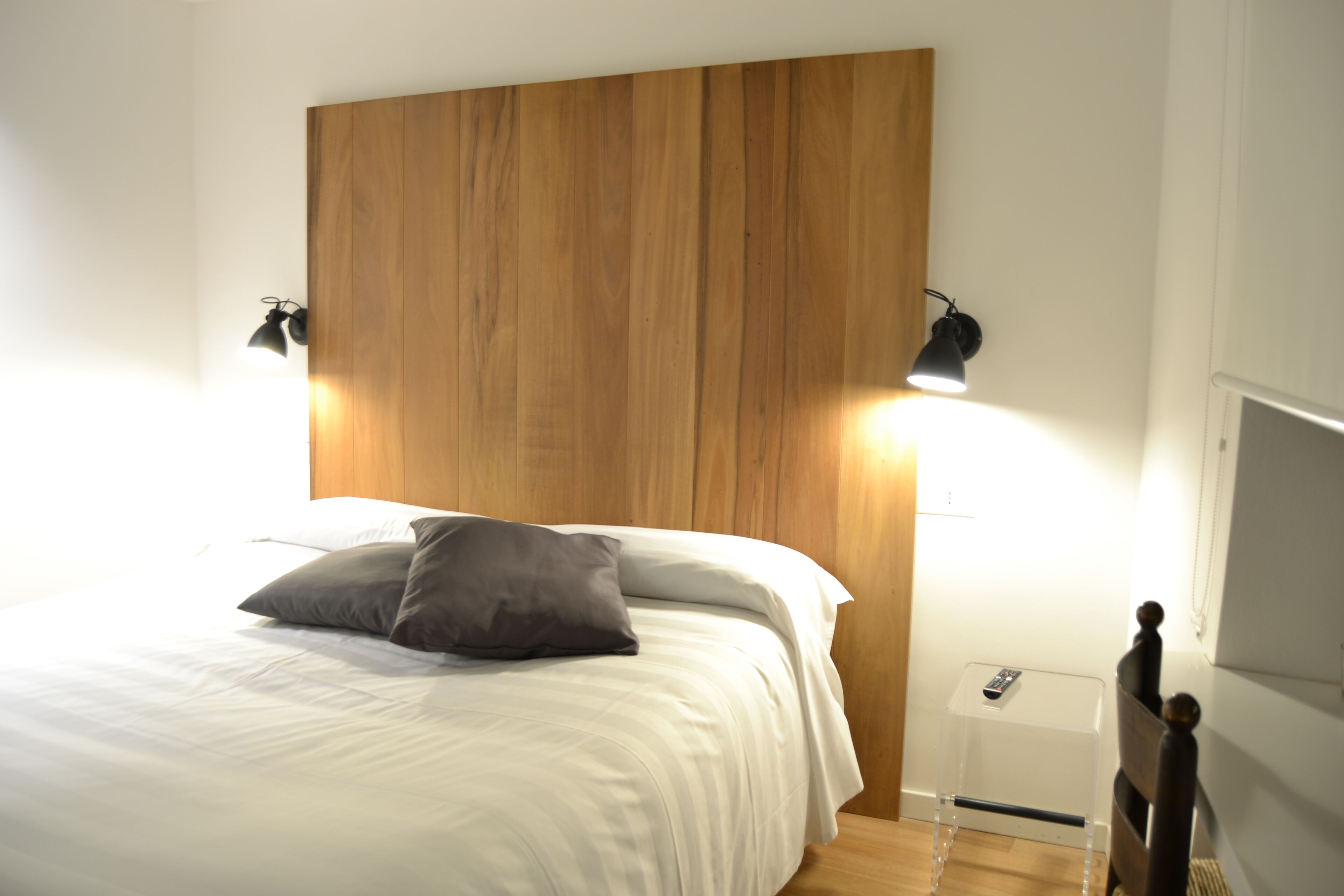 Cama bem arrumada com lençóis brancos e um único travesseiro cinza, cabeceira de madeira e duas luminárias fixadas na parede.