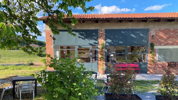 Un bâtiment à la façade en briques, doté de grandes fenêtres et d’un toit rouge. Il y a des espaces extérieurs aménagés avec des chaises et des tables en métal.
