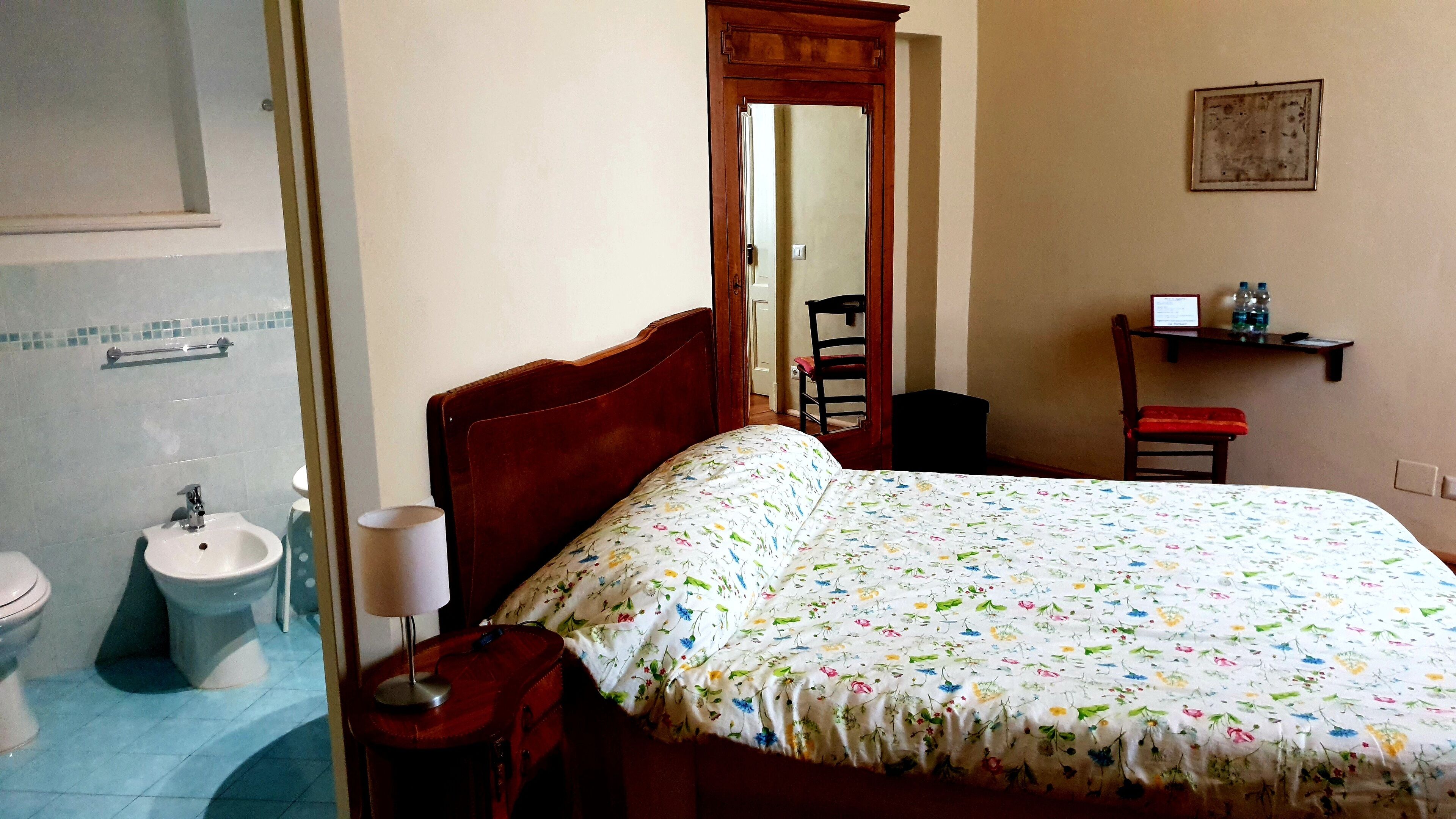A bedroom with a wooden bed, a bedside table with a lamp, a chair, a small shelf with toiletries, and a bathroom visible through an open door.