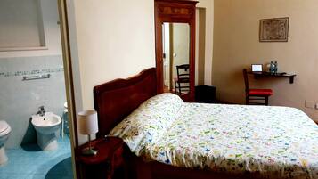 A bedroom with a wooden bed, a bedside table with a lamp, a chair, a small shelf with toiletries, and a bathroom visible through an open door.