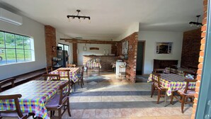 Un espace repas avec plusieurs tables et chaises, une cheminée en briques et une cuisine visible au fond.