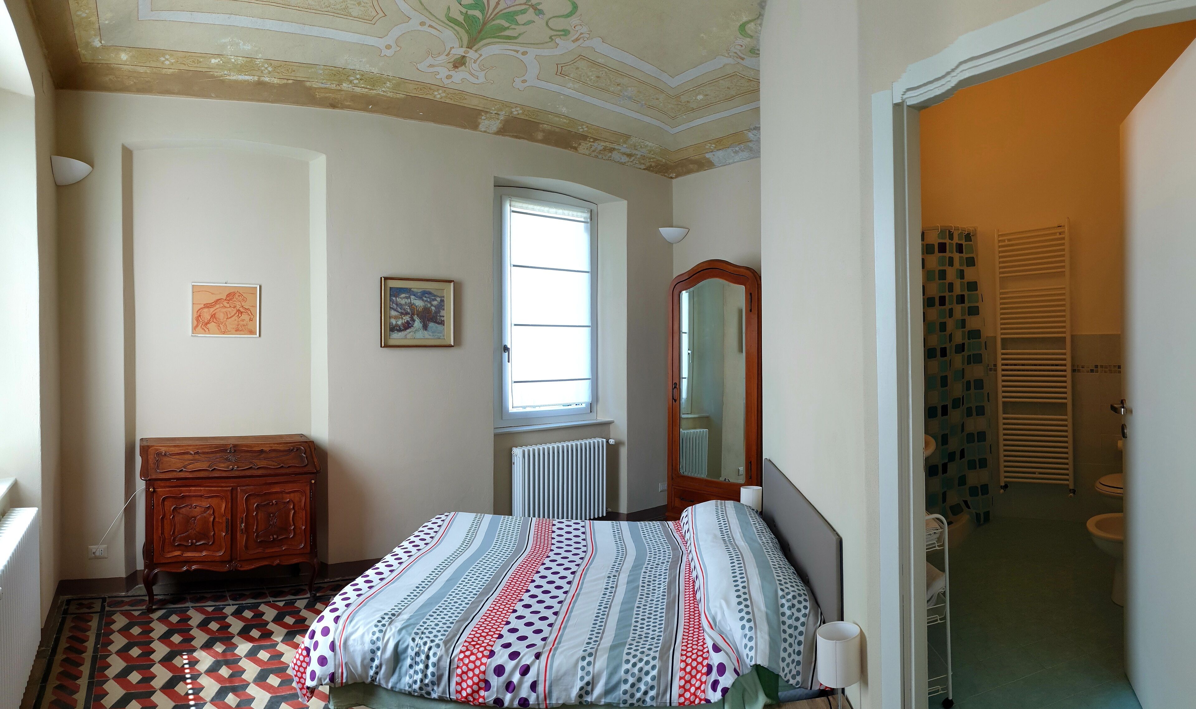 Un dormitorio con cama, cómoda, ventana, radiador y baño visible a través de una puerta abierta.