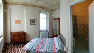 Un dormitorio con cama, cómoda, ventana, radiador y baño visible a través de una puerta abierta.