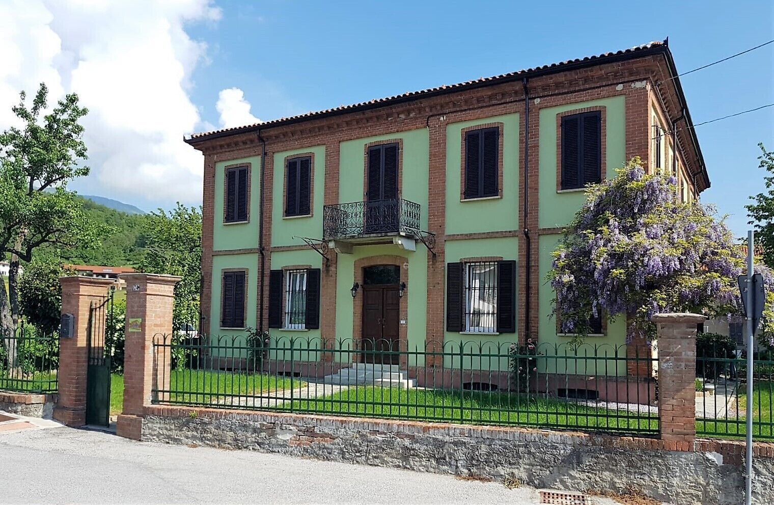 A two-story building with a green facade, multiple windows with shutters, a central entrance with a door, and a small balcony.