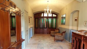 Un couloir orné de carreaux décoratifs, une porte en bois avec des panneaux de verre, un lustre et deux fauteuils.