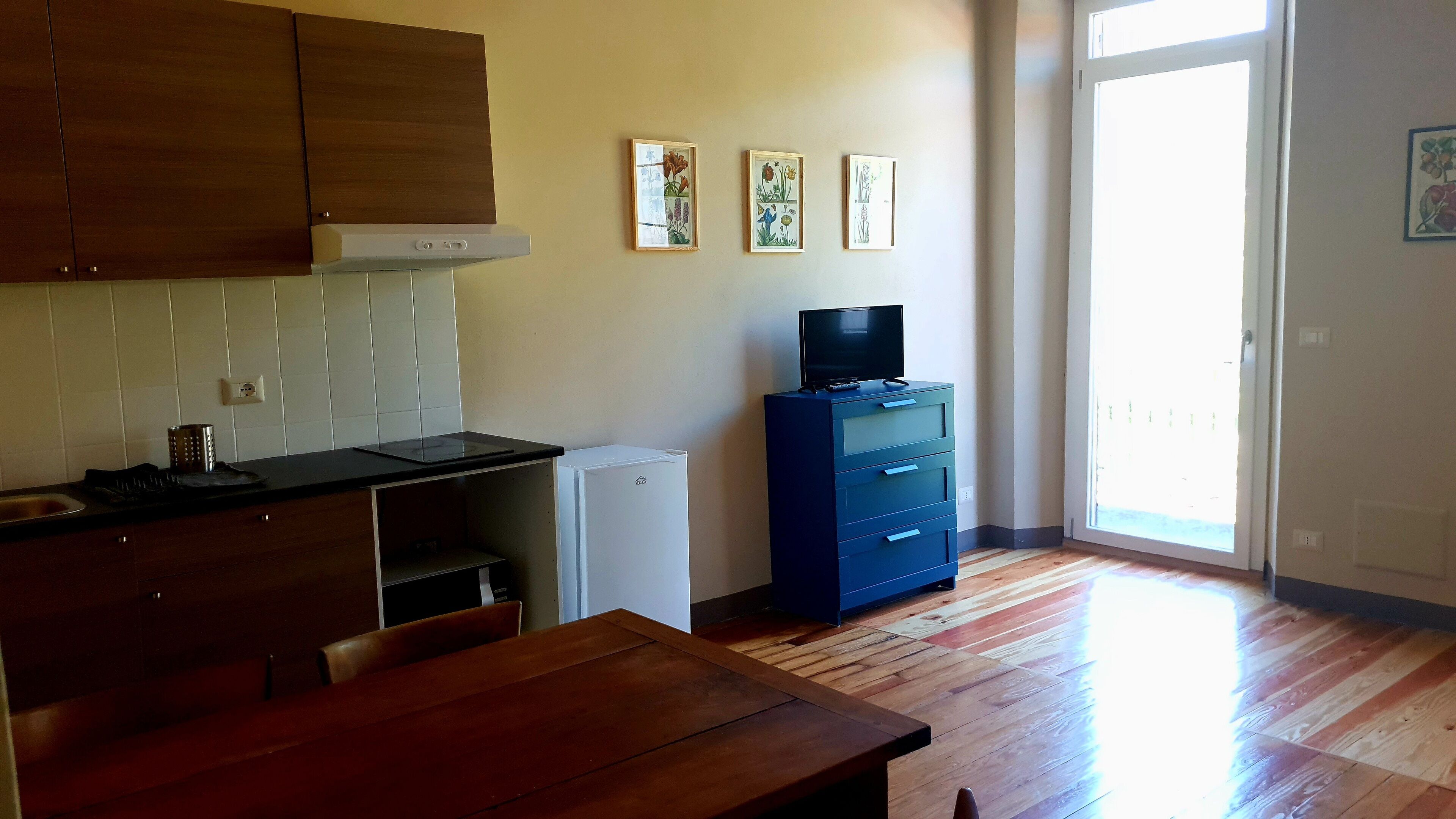 A kitchen with wooden cabinets, a white refrigerator, a black countertop, and a wooden dining table. There are framed botanical prints on the wall and a television on a blue dresser.