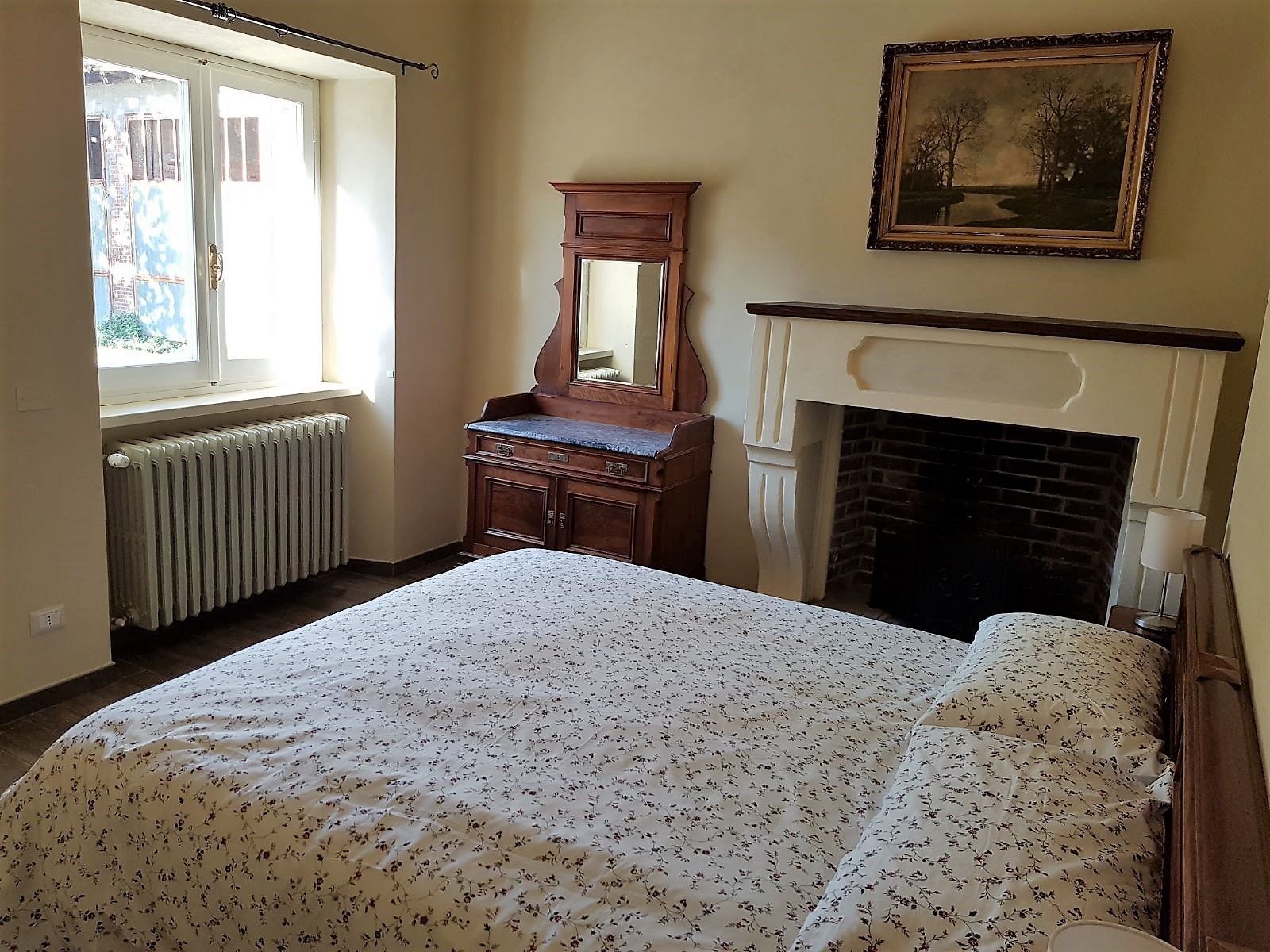 Ein Schlafzimmer mit Bett, Schminktisch, Spiegel, Kamin und einem Fenster mit Blick auf Gebäude.