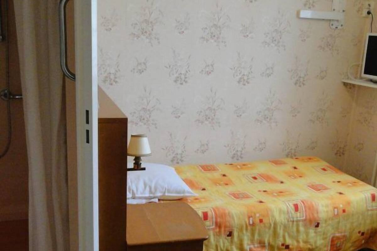 Quarto pequeno com uma cama, mesa de cabeceira, luminária e televisão em um suporte.