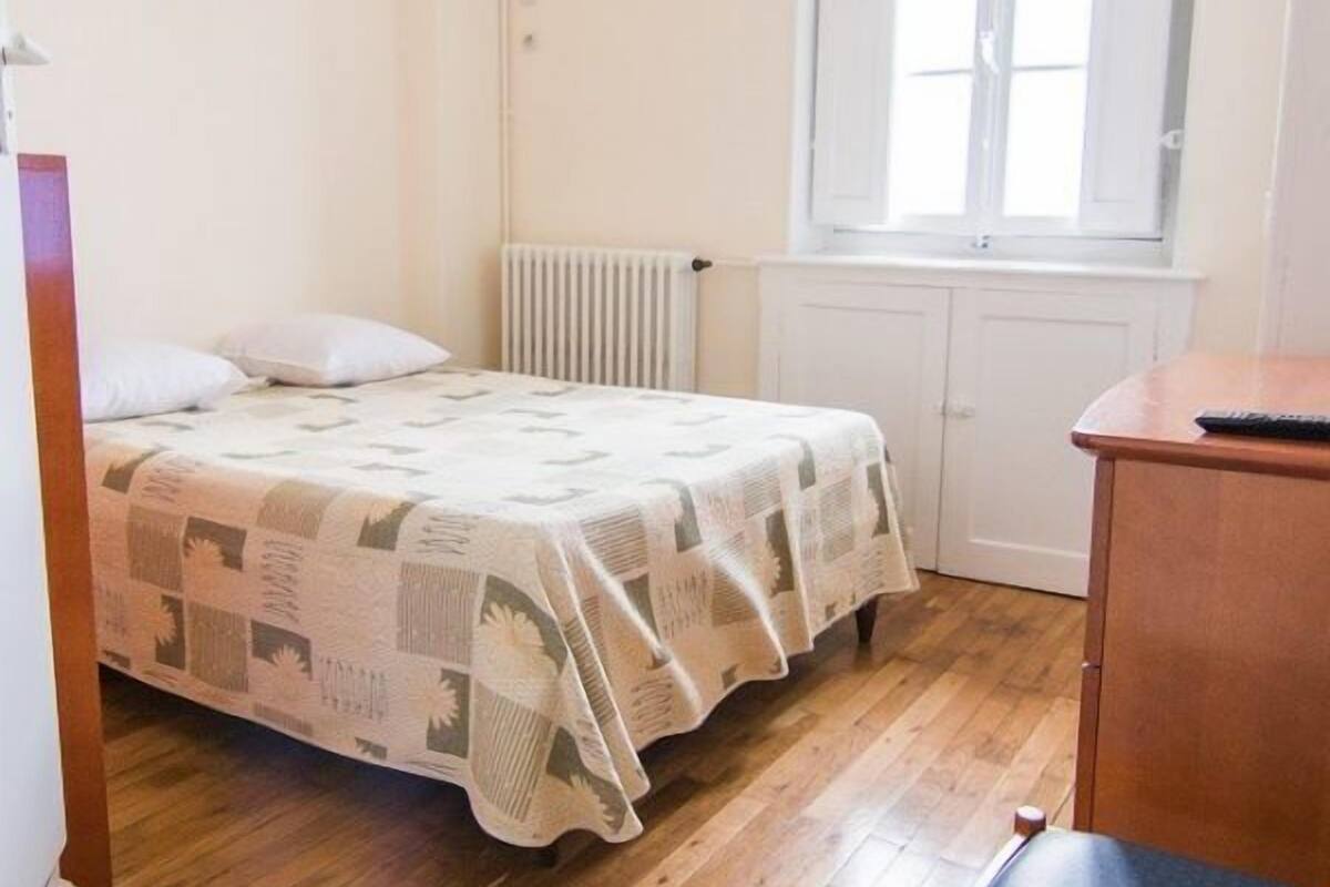 Quarto pequeno com cama de solteiro, piso de madeira, radiador, janela e escrivaninha com cadeira.