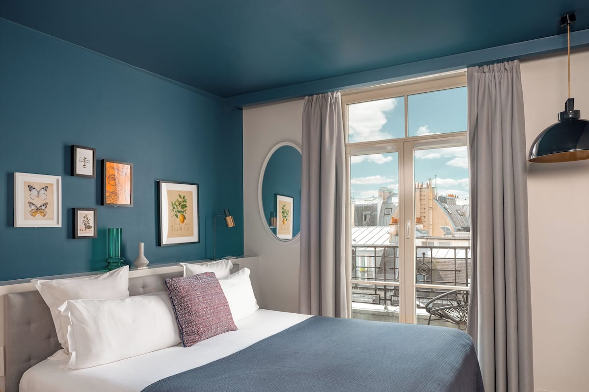 A bedroom with a bed, framed artwork, a mirror, and a view of buildings through a window.