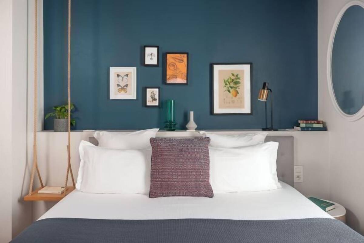 A bedroom with a bed, pillows, a swing, framed artwork, a lamp, and a mirror.