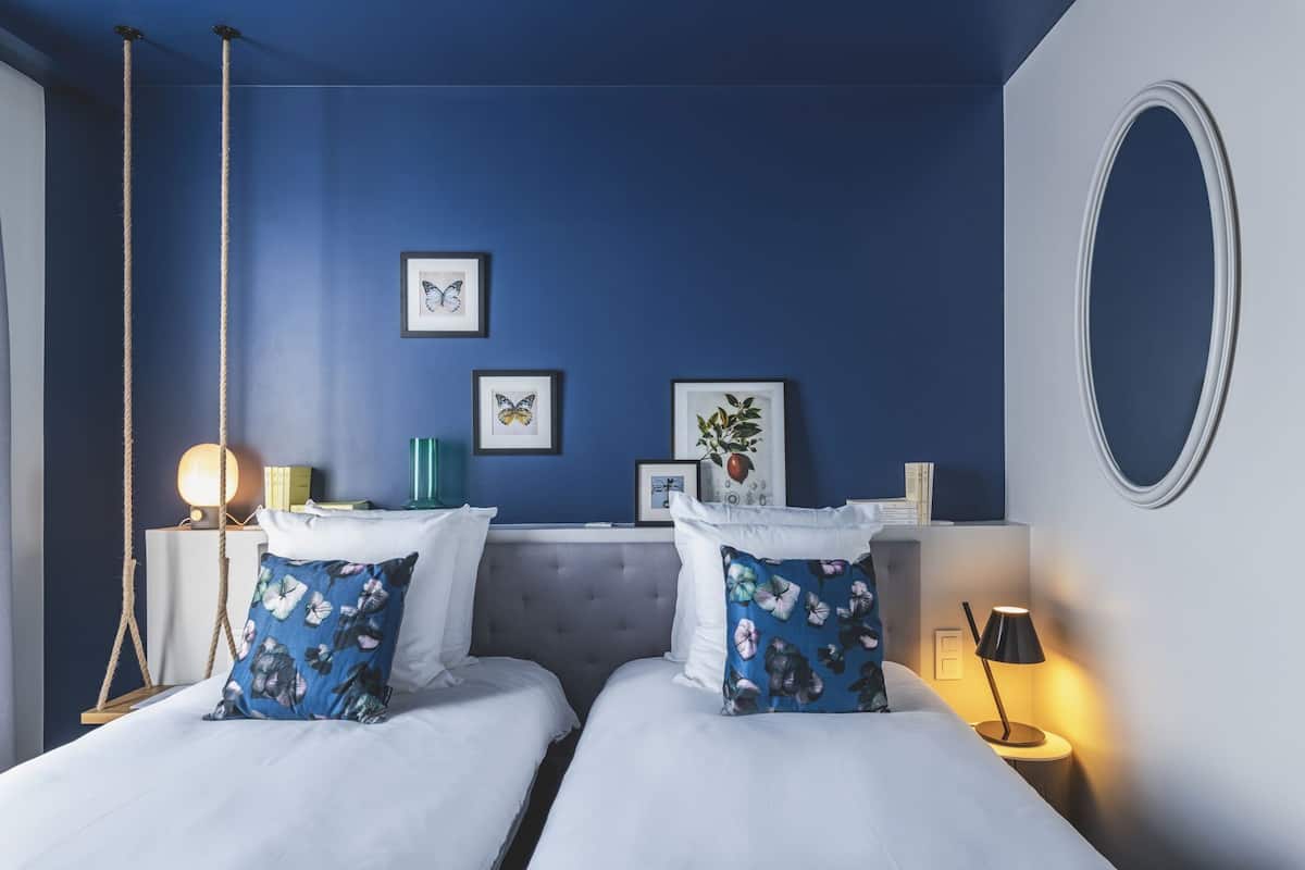 A hotel room with two beds, a blue wall, framed artwork, a mirror, and bedside lamps.