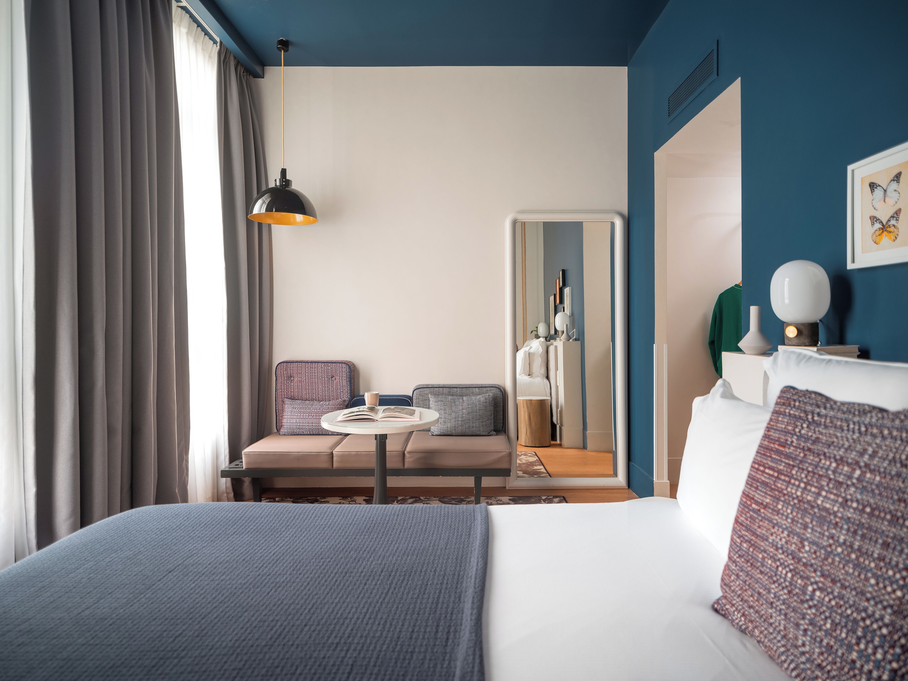 Habitación de hotel con una cama grande, una mesita redonda con un libro, una silla, un espejo y una mesita de noche con una lámpara.