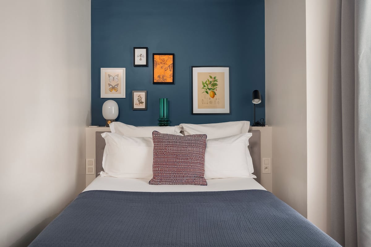 A neatly made bed with white linens and a blue blanket, a teal vase, and framed botanical prints on a dark blue wall.