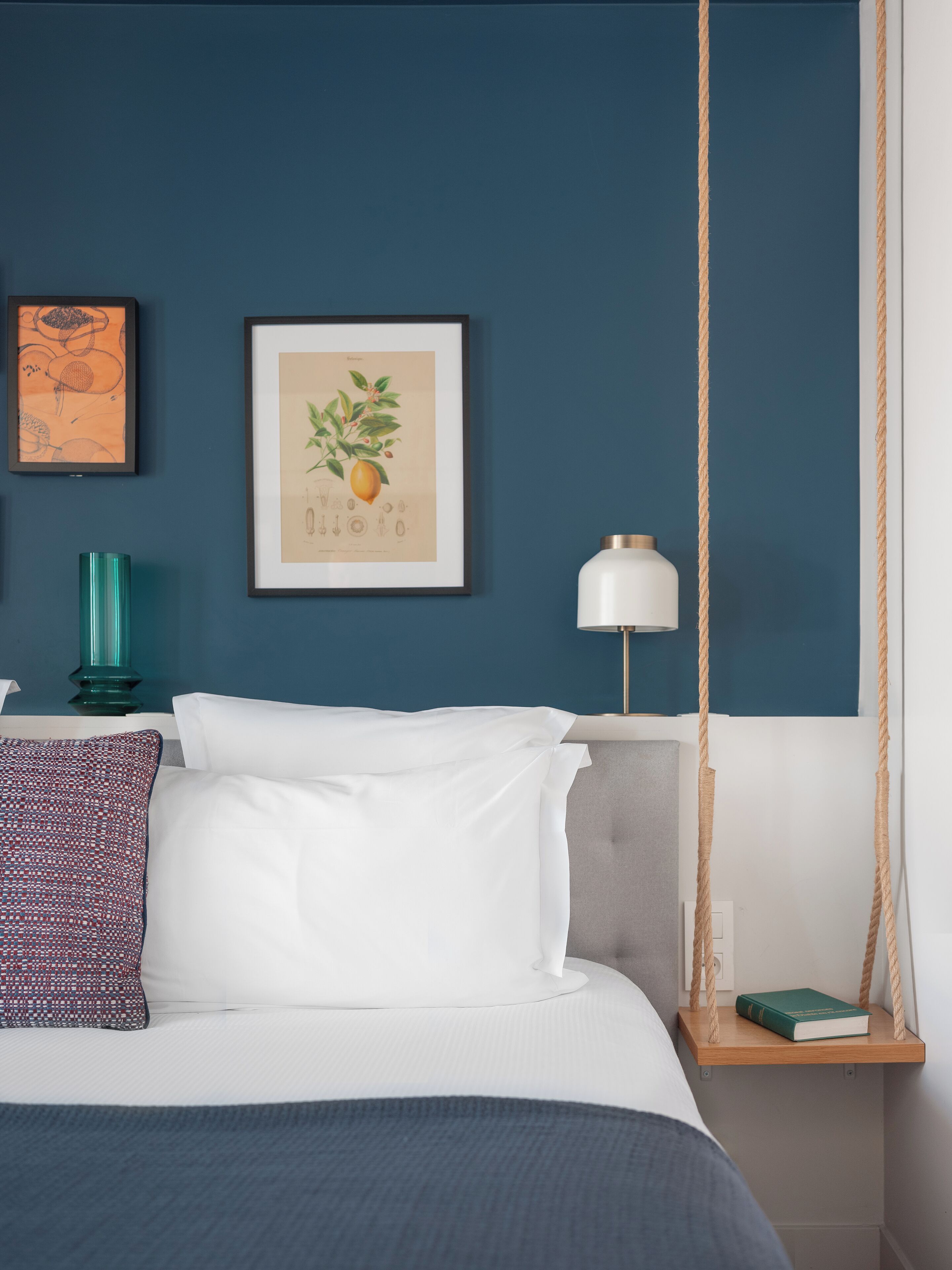 A neatly made bed with white and patterned pillows, a teal vase, and a framed botanical illustration on a dark blue wall.