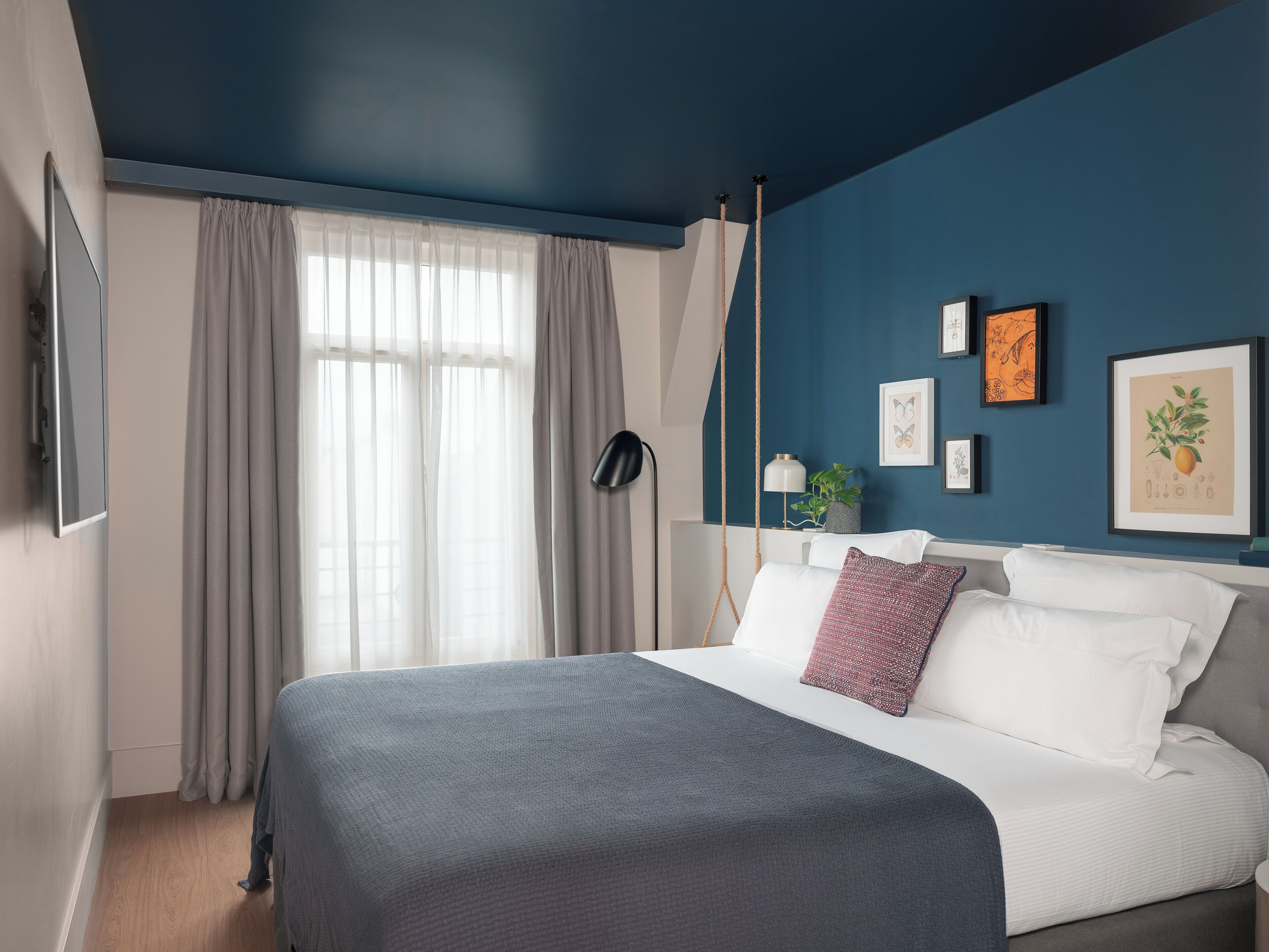 A modern bedroom with a large bed, a TV mounted on the wall, and a window with sheer curtains.