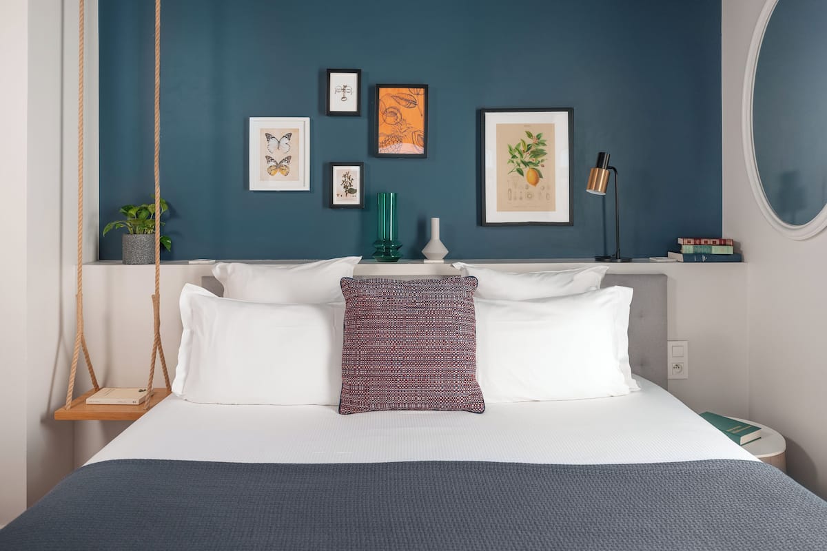 A bedroom with a bed, pillows, a hanging plant, framed artwork, and a bedside table with books and a lamp.