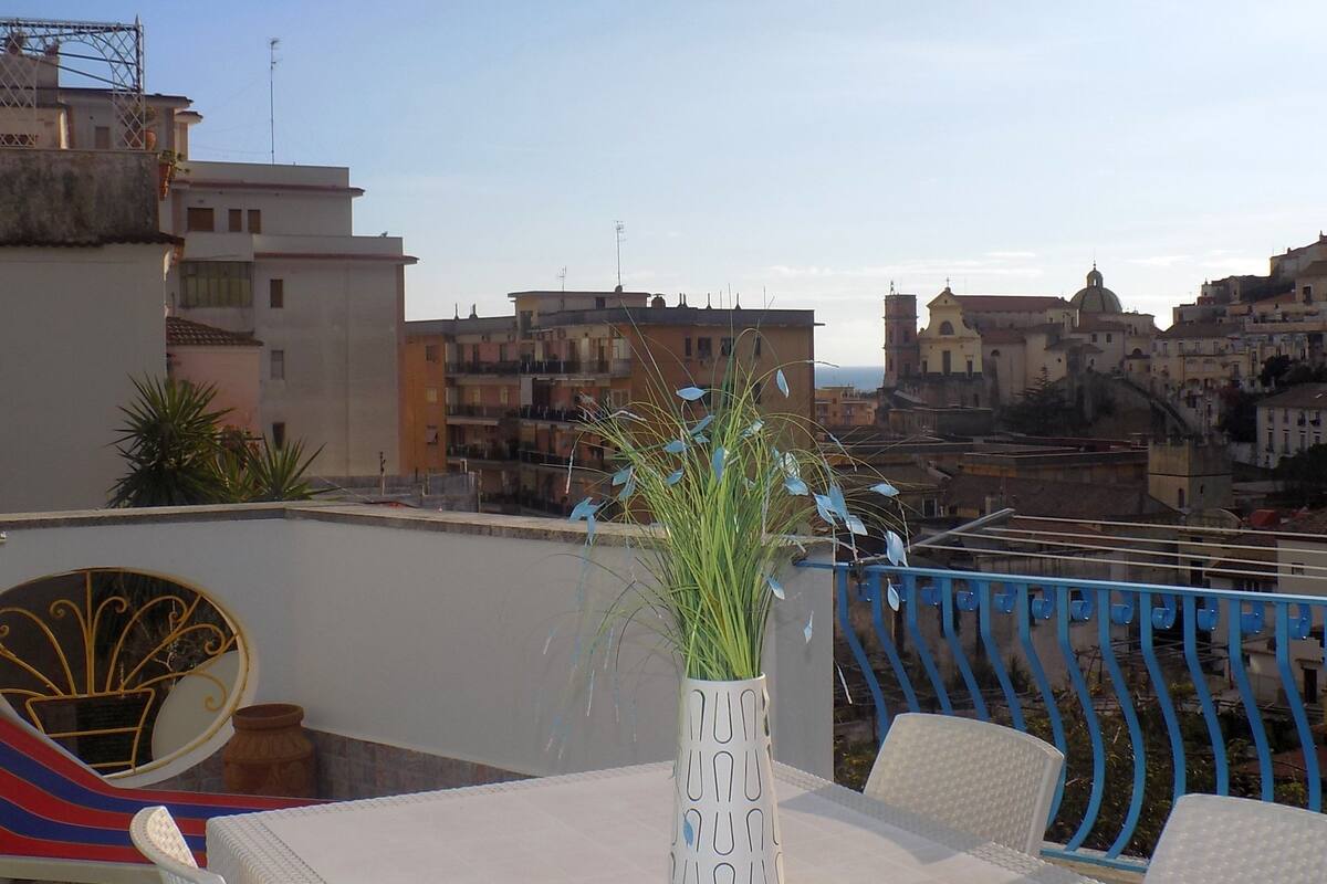 A rooftop terrace with a table, chairs, a decorative vase with plants, and a view of a cityscape with buildings and a dome structure.