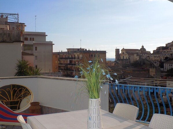 A rooftop terrace with a table, chairs, a decorative vase with plants, and a view of a cityscape with buildings and a dome structure.